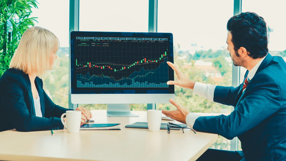 Deux analystes financiers en réunion discutant d'une analyse des indicateurs techniques affichée sur un grand écran d'ordinateur