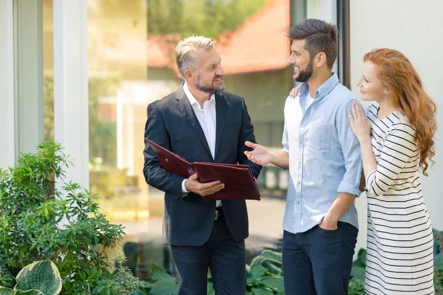 Un agent immobilier discute avec un jeune couple souriant lors d'une visite pour choisir la ville idéale.