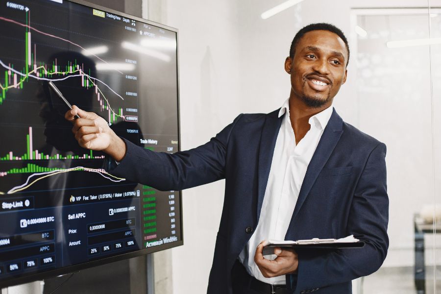 Un homme souriant en costume pointe du doigt des graphiques financiers affichés sur un grand écran dans un bureau moderne.